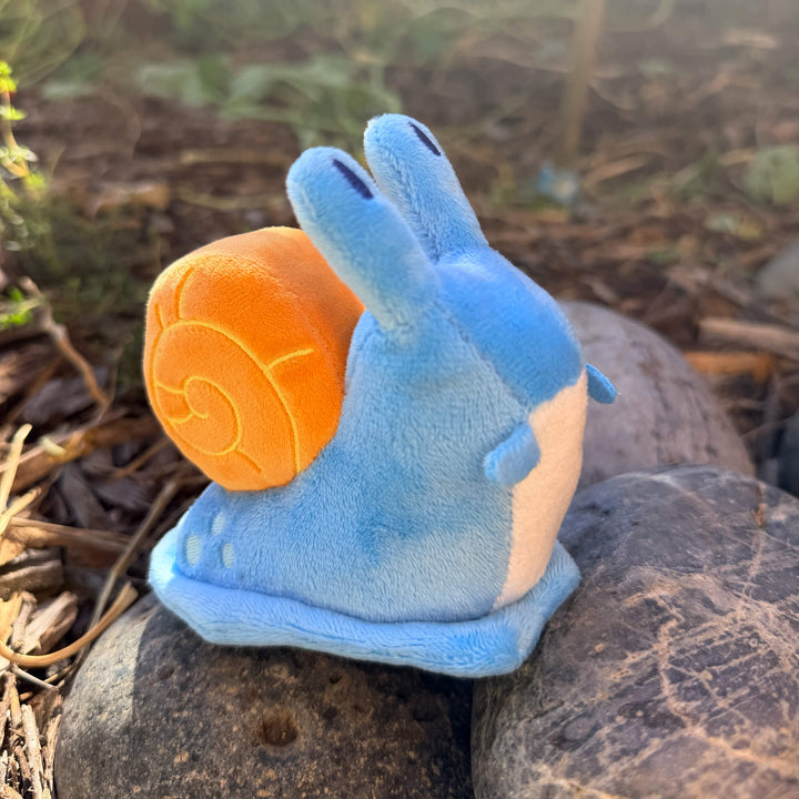 Blue plush snail toy keychain on a rock with a blurred natural background