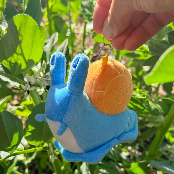 Blue plush toy snail keychain, held by a hand against a green leafy background.