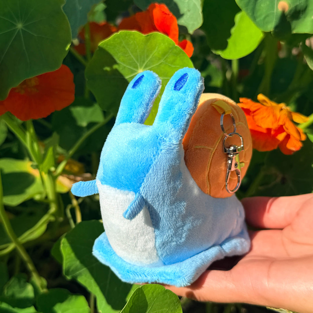 Blue plush toy snail with a keychain held in front of green plants and orange flowers
