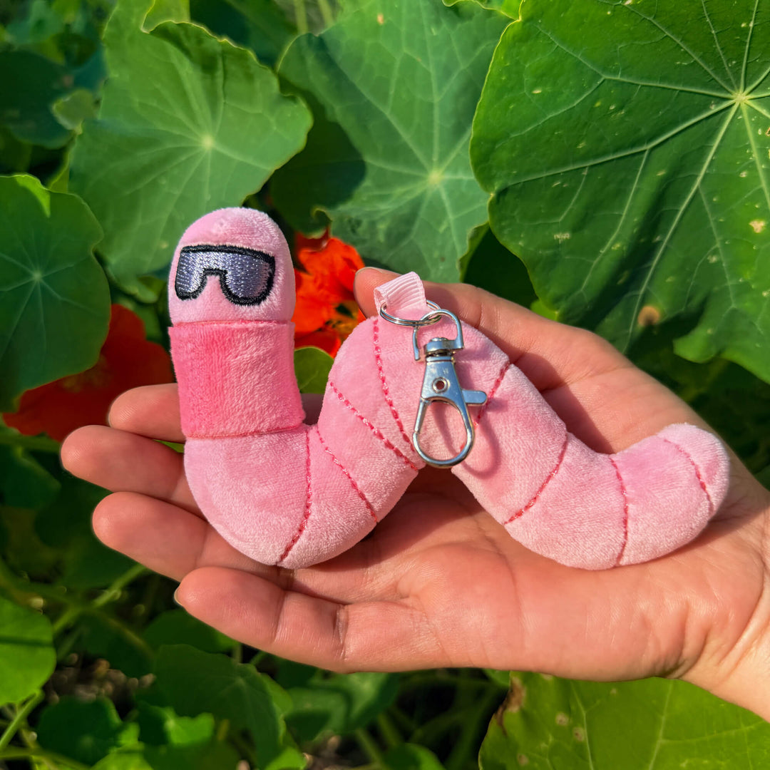 Pink plush toy earthworm keychain with sunglasses held in a hand against green leaves
