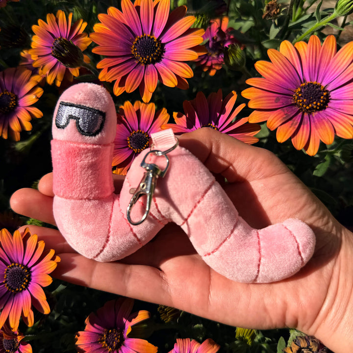 Pink plush earthworm toy keychain with sunglasses held in front of purple flowers