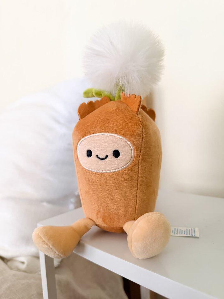 A cute plush toy shaped like a small orange-brown dandelion character with a white face patch, black embroidered eyes and smile, tan feet, and a fluffy white pom-pom dandelion seed on top, sitting on a white table against a soft cream background.