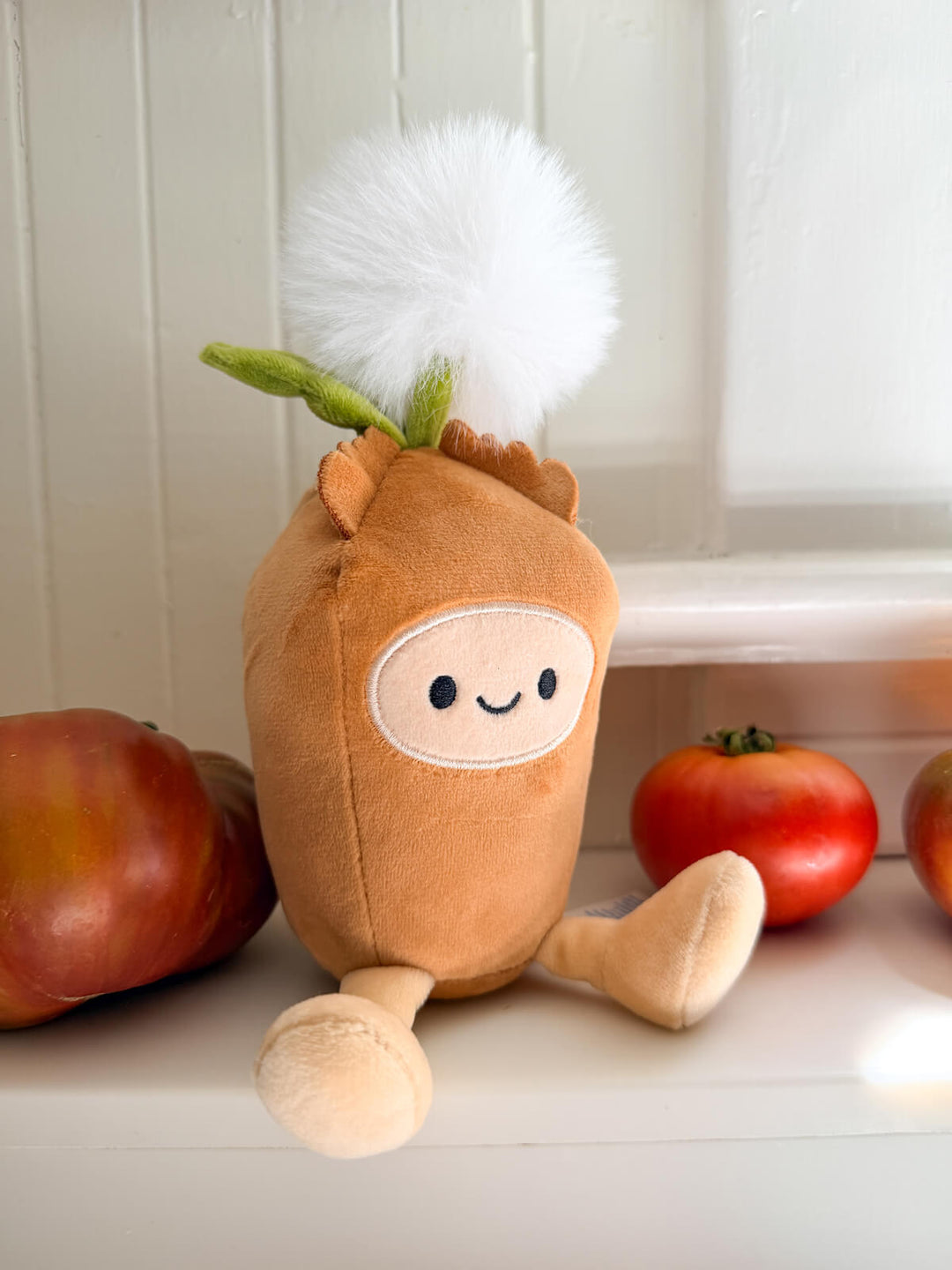 A plush dandelion toy in a warm brown fabric with a white fluffy pom-pom dandelion head on the top, styled on a white windowsill with red tomatoes in the background.