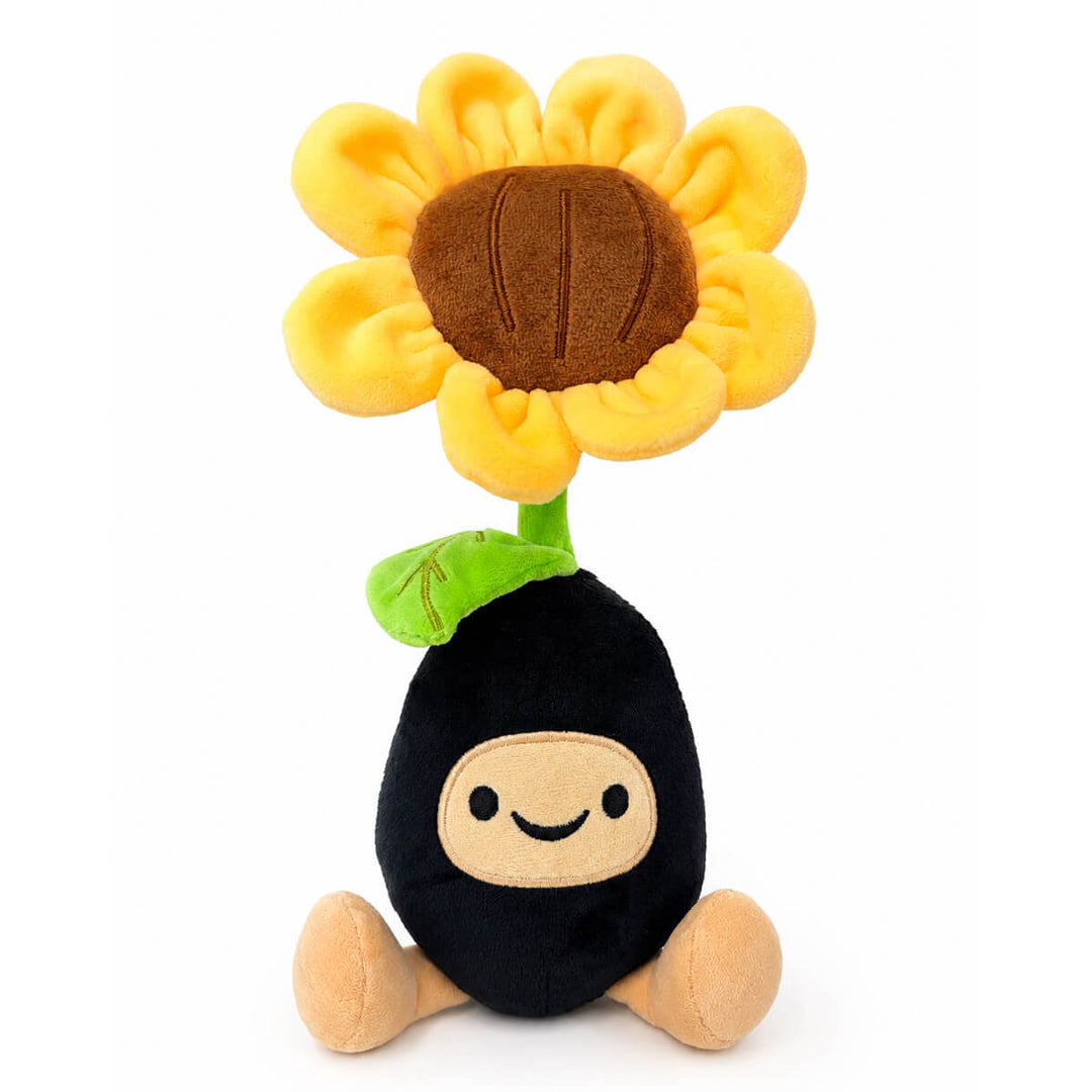 A plush toy shaped like a sunflower seed pod, featuring a black body, beige face and feet, a green stem, and a large yellow flower head with a brown center, set against a plain white background.